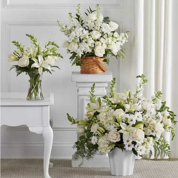 Set of three white floral arrangements with lilies and roses in glass vase, basket, and white container.