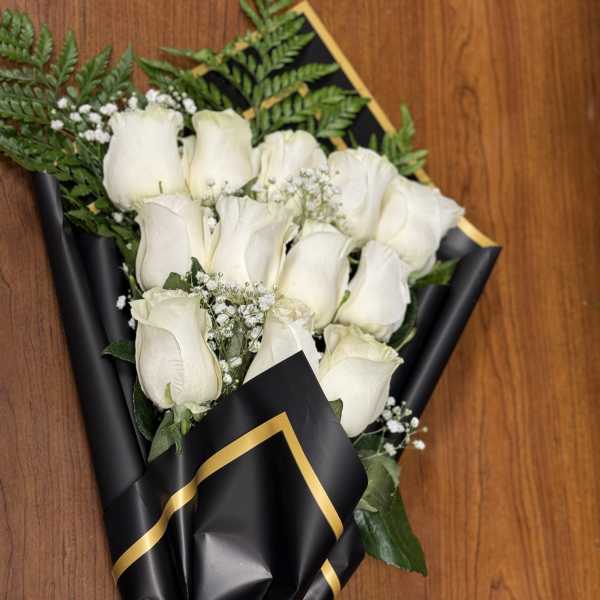 Wrapped bouquet of white roses with baby's breath in black paper with gold trim and white ribbon.