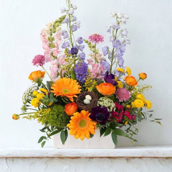 Colorful spring flower arrangement with orange gerberas and mixed blooms in a white box with a small bird nest