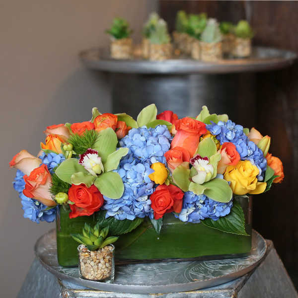 Rectangular floral arrangement with blue hydrangeas, orange and yellow roses, and orchids