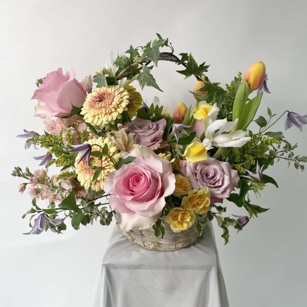 Pastel basket arrangement with pink roses, yellow gerberas, tulips, and an ivy-wrapped handle.