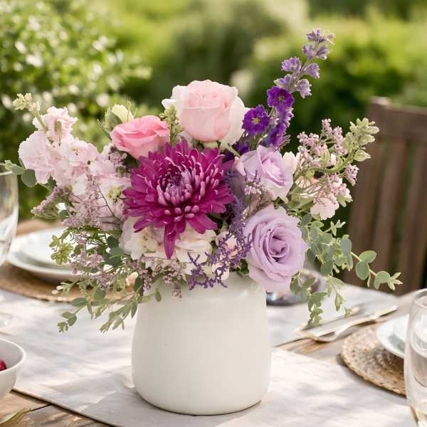 Pastel pink and lavender flower arrangement with a bold purple bloom in a white ceramic vase on a table.