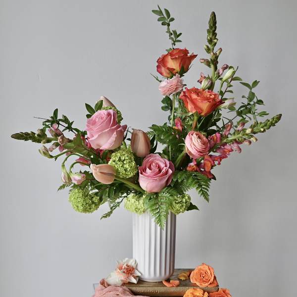 Pink and peach roses, tulips, and ranunculus in a tall white ribbed vase with green accents