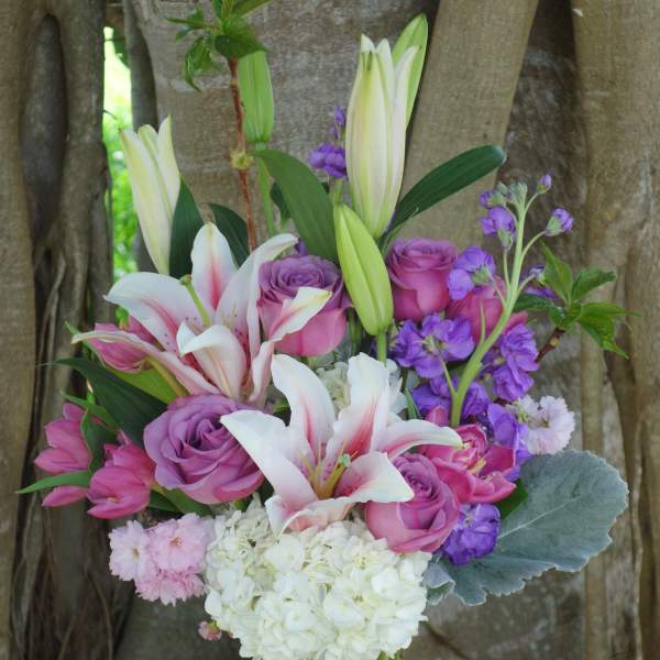 Tall glass vase of pink lilies, purple roses, white hydrangeas, and mixed pink and purple flowers.