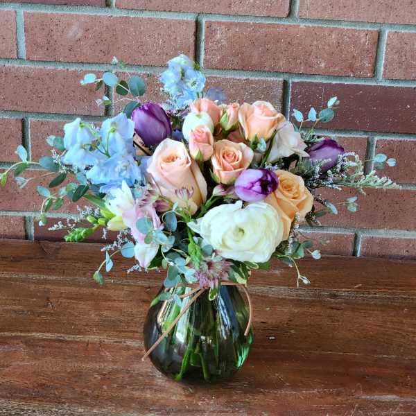 Peach roses, white ranunculus, and purple tulips arranged in a round green glass vase.