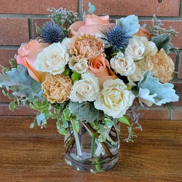 Peach and cream roses with blue thistle-style blooms arranged in a clear glass vase