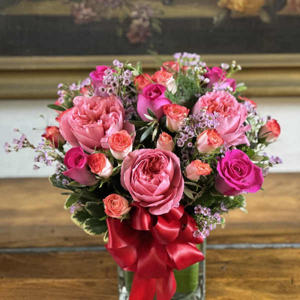 Compact arrangement of pink and hot pink roses in a clear glass vase with a red ribbon bow.