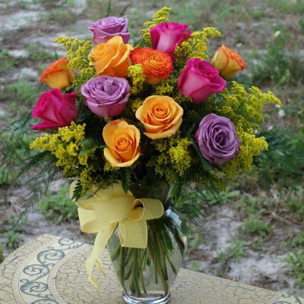 Mixed bouquet of yellow, orange, pink, and lavender roses in a clear vase with a yellow bow