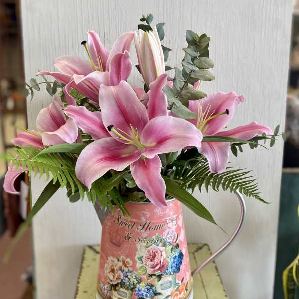 Pink lilies arranged in a decorative metal pitcher with greenery