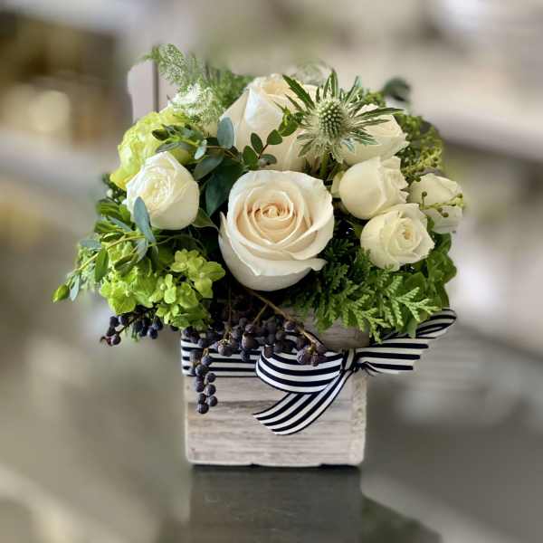 White roses and green flowers arranged in a small wooden box with a striped ribbon