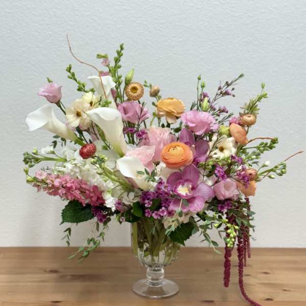 Asymmetrical pastel arrangement with calla lilies, ranunculus, orchids, and mixed blooms in a clear glass vase