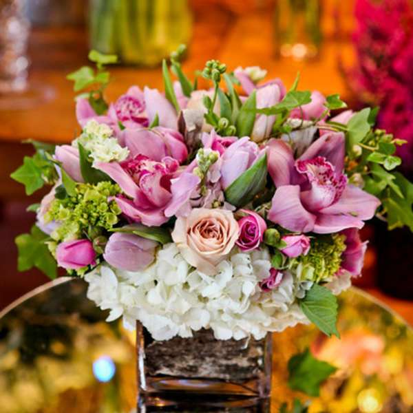 Low cube vase arrangement of pink orchids, roses, and white hydrangeas on a mirrored base.