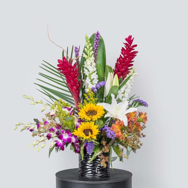 Tropical flower arrangement with sunflowers, lilies, orchids, and red ginger in a black vase