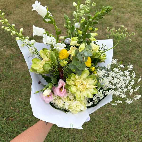Handheld bouquet of yellow, white, and pink mixed flowers wrapped in white paper over a grassy lawn