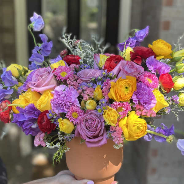 Colorful bouquet of roses, ranunculus, and purple blooms in a tan vase