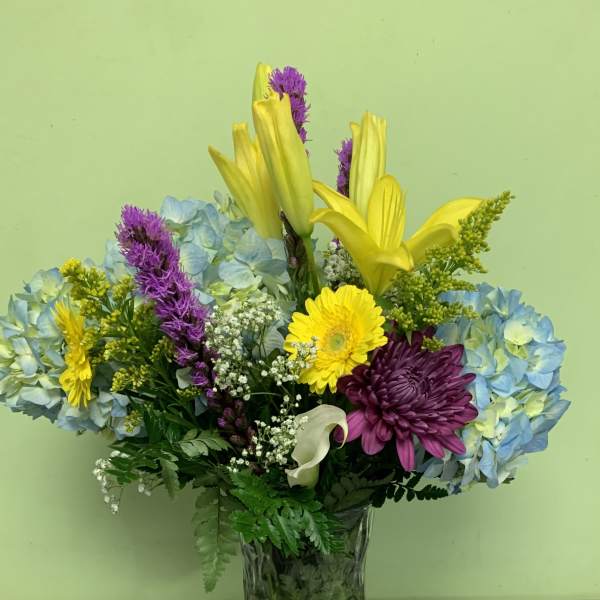 Mixed bouquet in a glass vase with blue hydrangeas, yellow lilies, and purple chrysanthemums