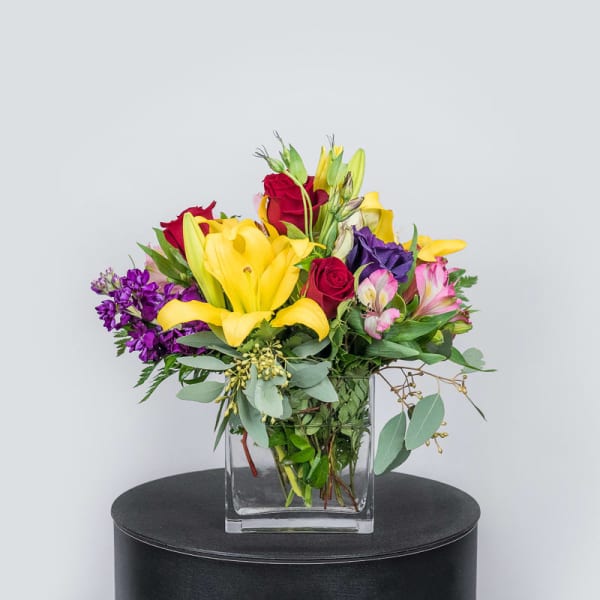 Mixed bouquet of yellow lilies, red roses, and purple flowers in a glass vase