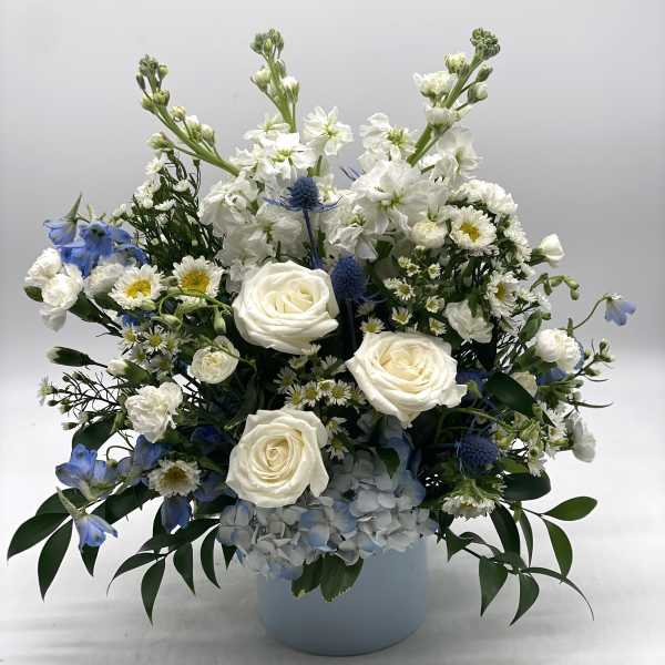 White and blue arrangement with roses, hydrangeas, and daisies in a light blue cylinder vase