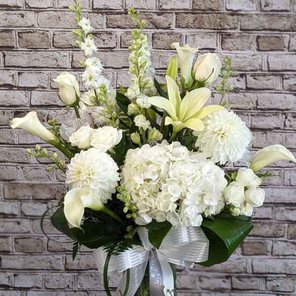Tall white floral arrangement with lilies, roses, hydrangeas and calla lilies in a glass vase with white ribbon