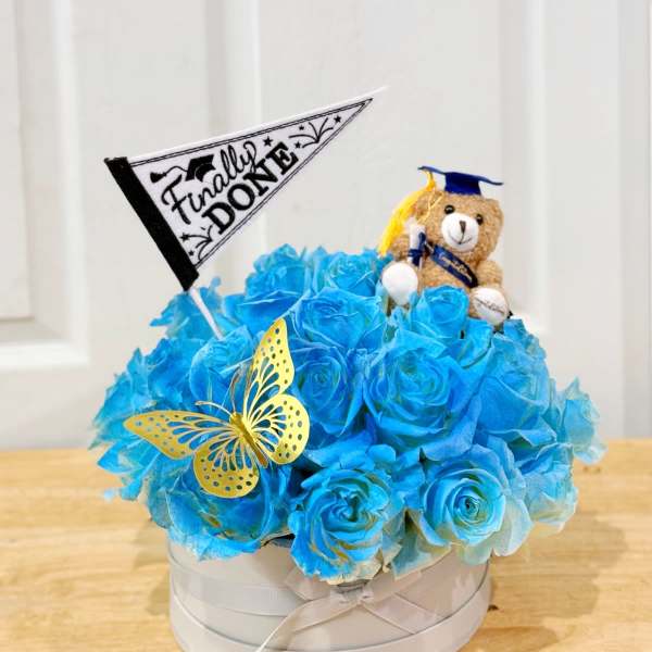 Blue rose arrangement in a round box with a teddy bear and graduation flag
