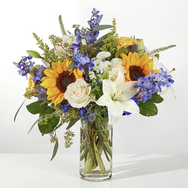 Bouquet of sunflowers, white lilies, and blue flowers in a clear glass vase