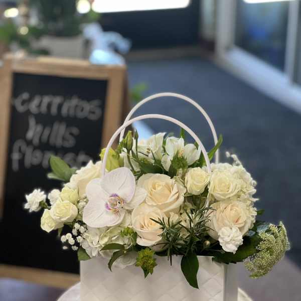 White floral arrangement in a handled gift box with roses and orchids
