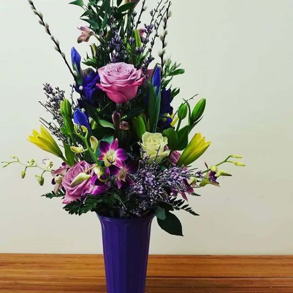 Tall mixed bouquet in a purple vase with pink, yellow, and blue flowers
