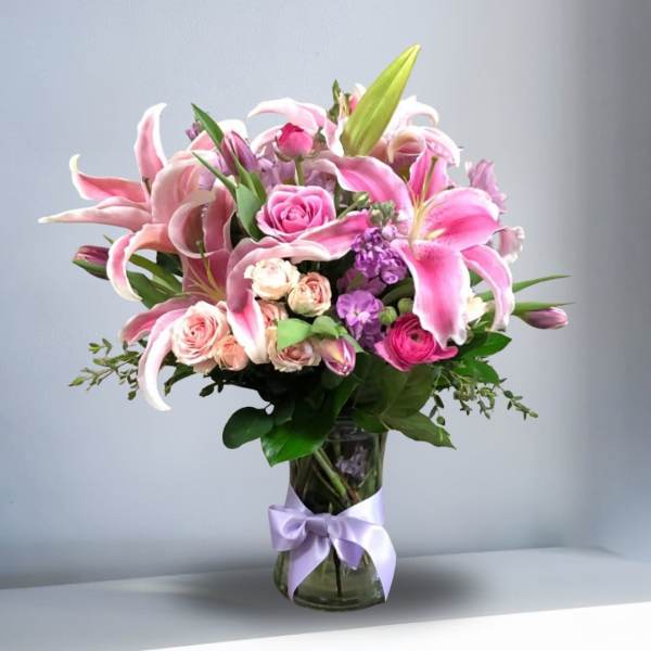 Pink lilies, roses, and purple flowers in a clear glass vase with a lavender ribbon.