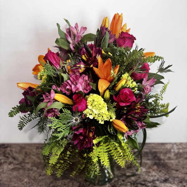 Mixed bouquet of pink roses, orange lilies, and purple flowers in a glass vase