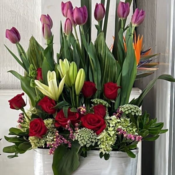 Tall floral arrangement with red roses, purple tulips, and white lilies in a white container