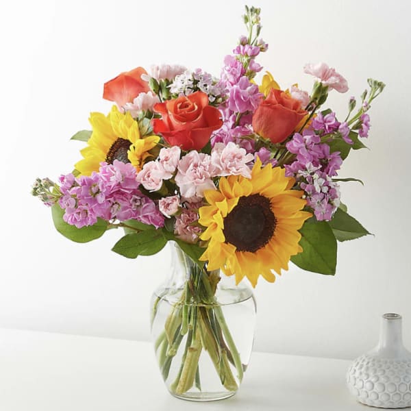 Mixed bouquet of sunflowers, orange roses, and pink blooms arranged in a clear glass vase.