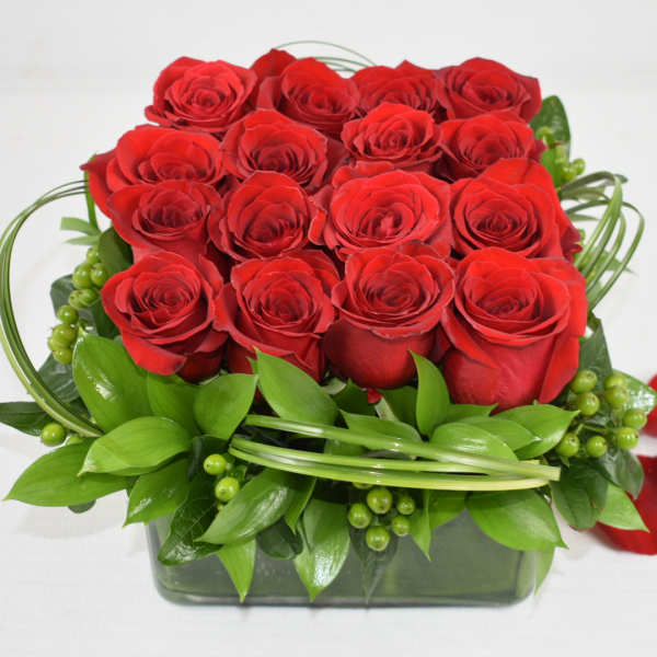 Square arrangement of red roses with decorative green loops in a glass cube vase