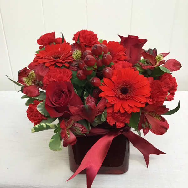Compact arrangement of red roses, gerbera daisies, carnations, and alstroemeria in a red cube vase with ribbon.