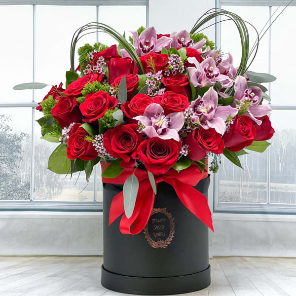 Red roses and pink orchids arranged in a black hatbox with a red ribbon