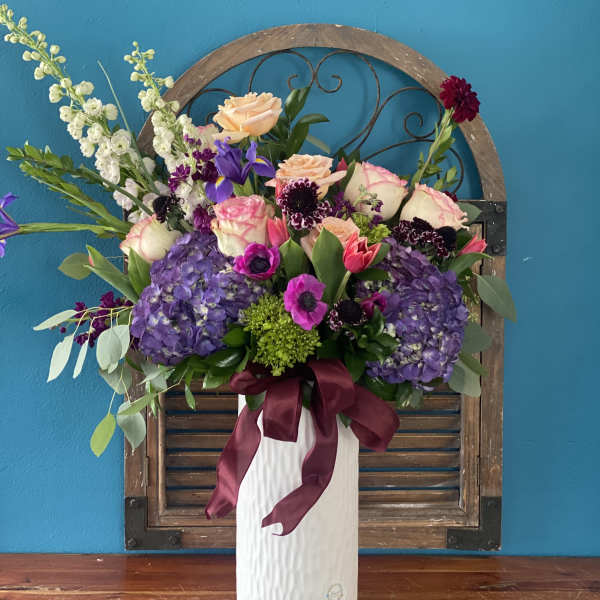 Tall floral arrangement in a white vase with purple, pink, and peach blooms