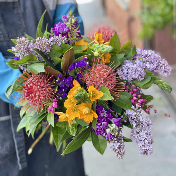 Handheld bouquet with yellow blooms, purple flowers, and pink pincushion protea
