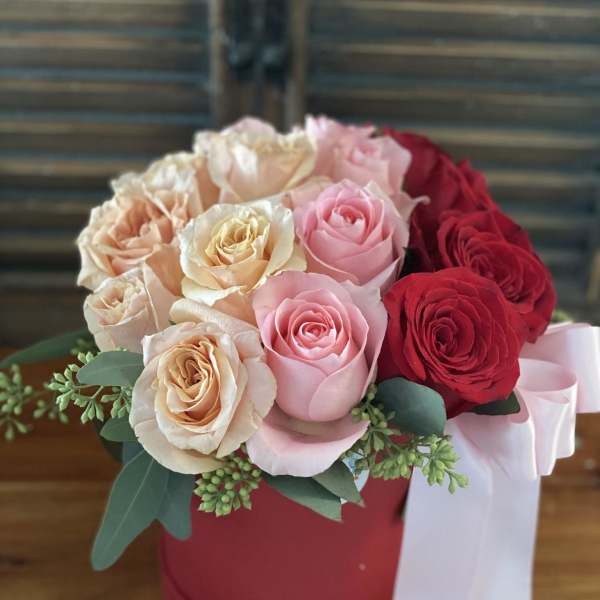 Bouquet of pink, peach, and red roses in a red hat box with a white ribbon
