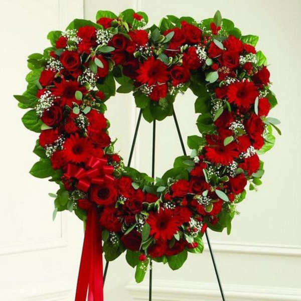 Heart-shaped standing wreath of red roses and other red flowers with a red ribbon bow.