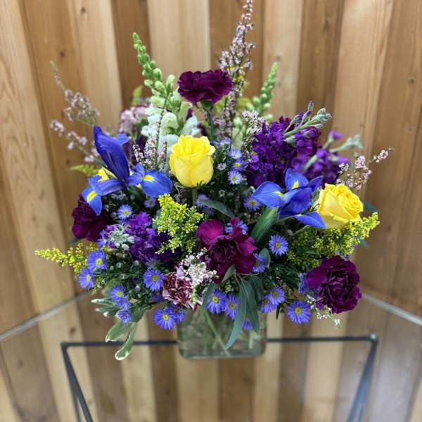 Mixed bouquet of yellow roses, blue irises, and purple flowers in a glass vase