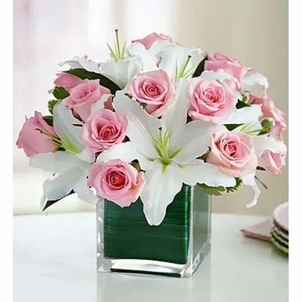 Arrangement of pink roses and white lilies in a clear cube vase lined with green.