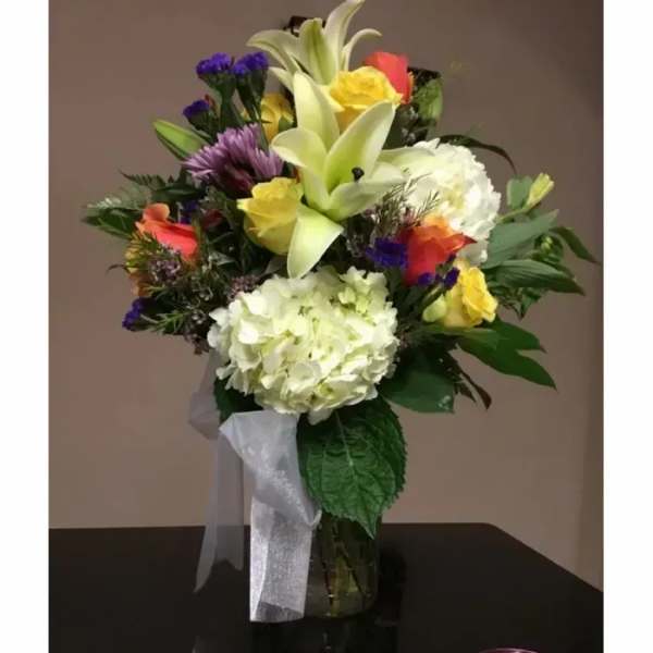 Mixed bouquet of white hydrangeas, lilies, yellow and orange roses in a clear glass vase with ribbon