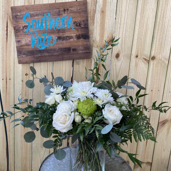 White roses and mums with a green accent bloom in a clear glass vase on a metal table.
