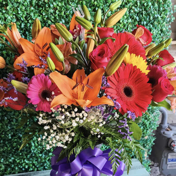 Bright mixed bouquet with lilies, gerbera daisies, roses, and a purple ribbon in a glass vase