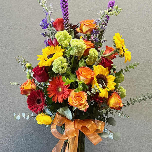 Mixed bouquet with roses, sunflowers, gerbera daisies, and purple spikes in a glass vase