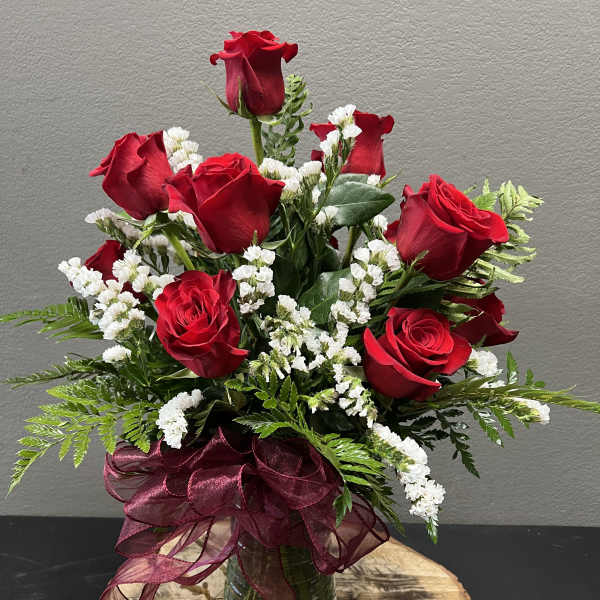 Red roses arranged in a glass vase with white filler flowers and a burgundy ribbon