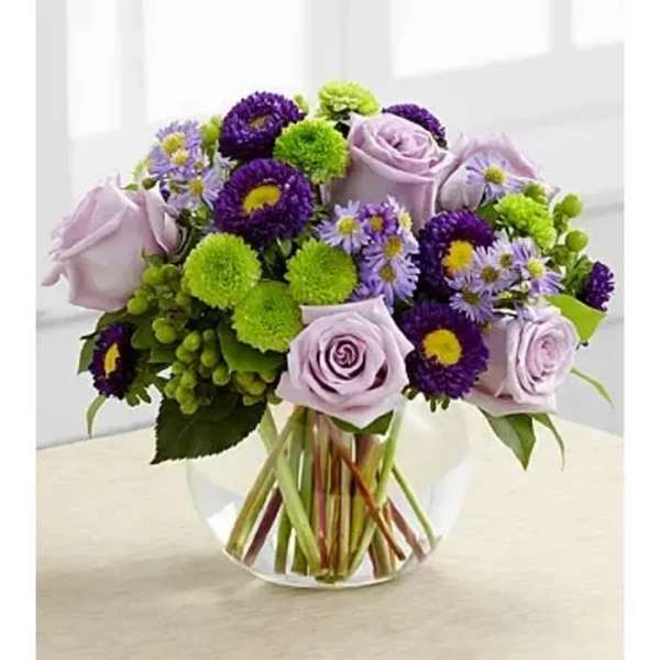 Round arrangement of lavender roses, purple asters, and green button mums in a clear glass bowl vase