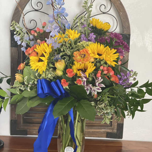 Mixed bouquet with sunflowers, purple blooms, and a blue ribbon in a glass vase