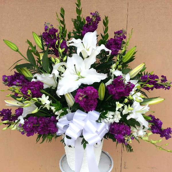 White lilies and purple flowers arranged in a white vase with a bow.