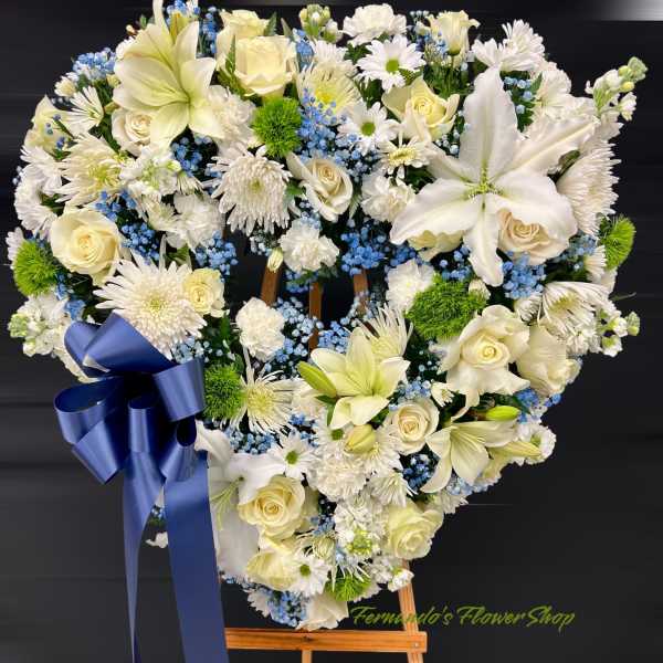 Heart-shaped white and blue standing wreath with lilies, roses, and a navy bow on an easel
