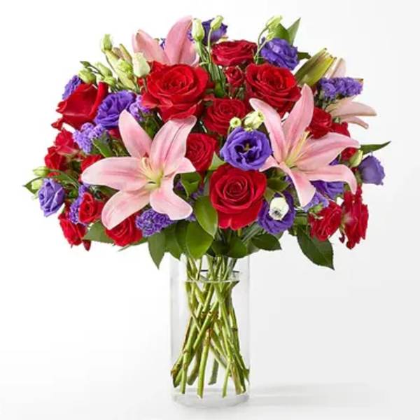Vase arrangement of pink lilies, red roses, and purple blooms in a clear glass cylinder